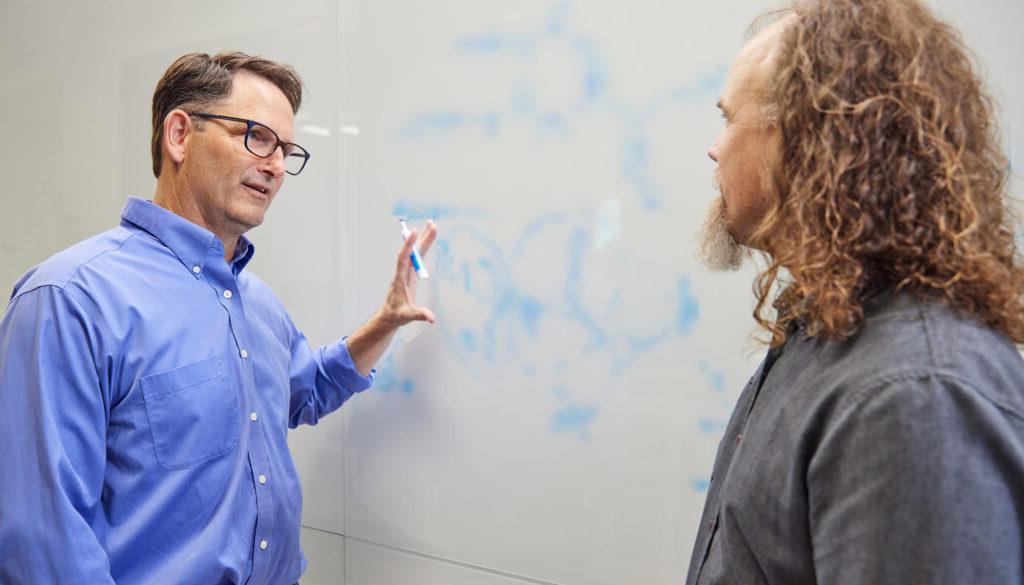 博彩平台网址大全 scientists Brett Crawford and Dan Wendt talking by a whiteboard with a scientific formula